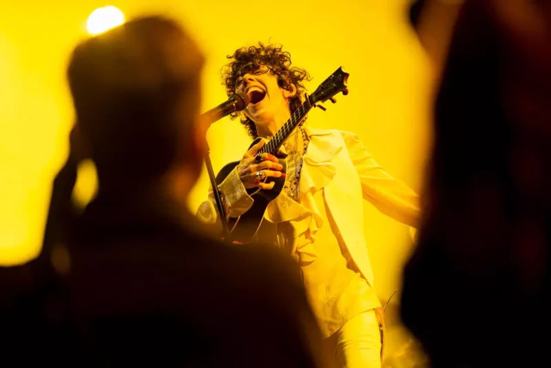 Interpret vášnivo spieva do mikrofónu a drží gitaru na pódiu. Jasné žlté osvetlenie vytvára energickú a dynamickú atmosféru.