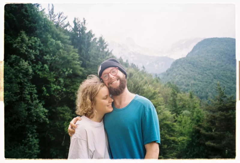 Usmievavý pár sa objíma v bujnom zelenom lese s hmlistými horami v pozadí. Scéna vyžaruje teplo, pokoj a prírodnú krásu.