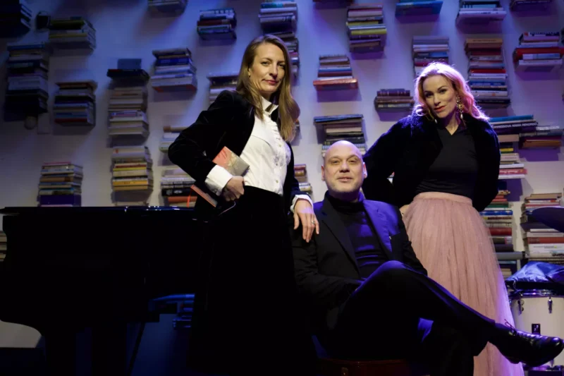 Traja ľudia sebavedomo pózujú v slabo osvetlenej miestnosti s policami na knihy ako pozadím. Dve ženy stoja a muž sedí na stoličke. Čiastočne je viditeľný klavír.