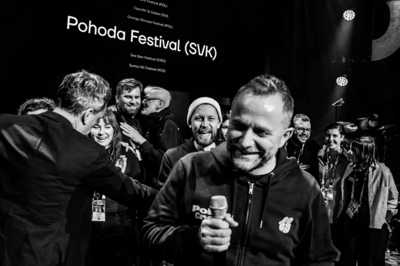 Skupina ľudí oslavuje na pódiu na festivale Pohoda. Muž drží mikrofón a usmieva sa. V pozadí sú zobrazené názvy festivalov. Slávnostná a radostná atmosféra.