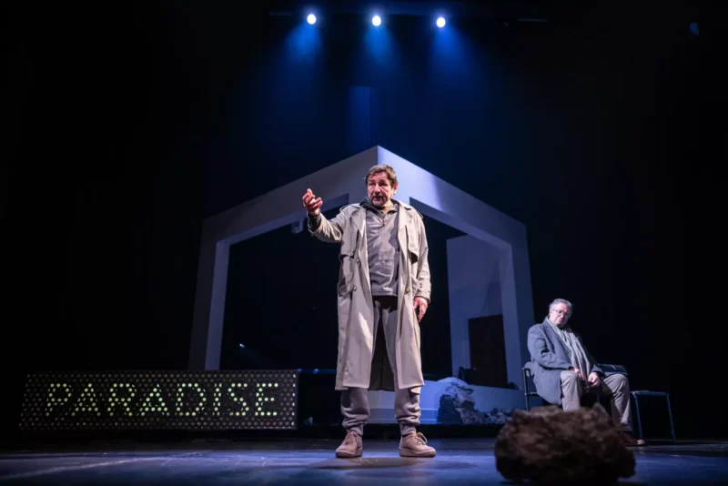 Muž v trenčkote vášnivo gestikuluje na pódiu pod dramatickým modrým osvetlením. Na nápise je „RAJ“. V pozadí sedí ďalší muž a pozoruje.