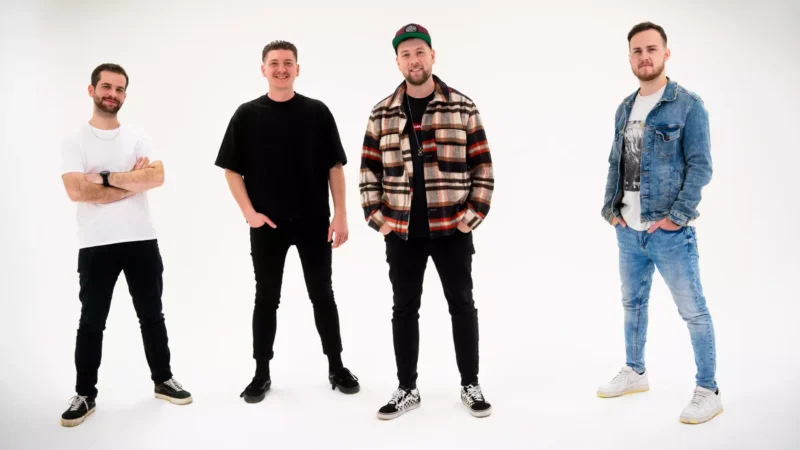 Štyria muži stoja sebavedomo na bielom pozadí. Majú na sebe ležérne oblečenie: tričká, džínsy a tenisky. Nálada je uvoľnená a priateľská.