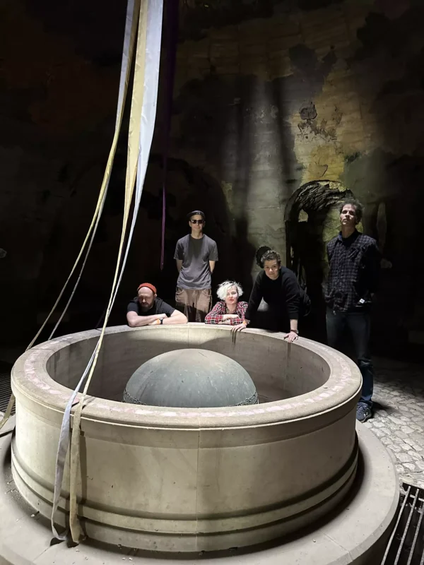 Skupina piatich ľudí stojí okolo kruhovej kamennej stavby so stuhami visiacimi zhora v slabo osvetlenom priestore podobnom jaskyni, čo vyvoláva zvedavosť a intrigu.