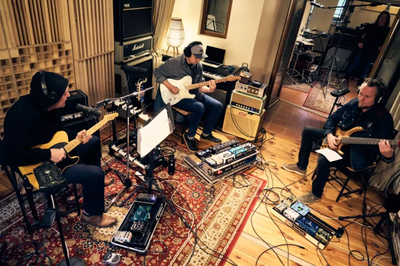 Traja hudobníci hrajú na elektrické gitary a basgitaru v útulnom štúdiu s teplým osvetlením. Miestnosť je plná káblov a zosilňovačov, čo vytvára kreatívnu atmosféru.