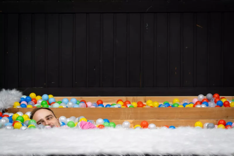 Osoba hravo ponorená v farebnej jame s loptičkami, pričom je viditeľná iba jej tvár. Pozadie tvorí tmavá drevená stena, ktorá vytvára radostný kontrast.