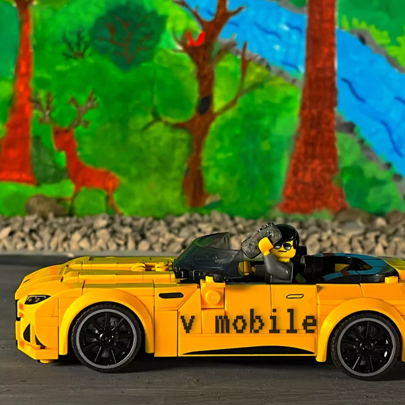 Žltý kabriolet LEGO s nápisom „v mobile“ na boku a figúrkou telefonujúcou jazdí po sivom povrchu. Pozadie: svetlá nástenná maľba so stromami a riekou.