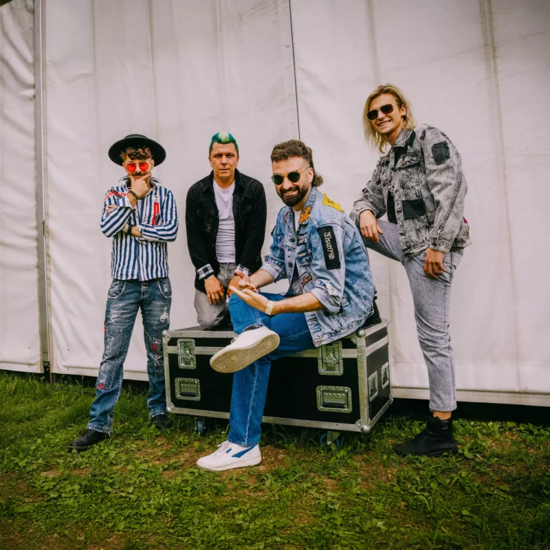 Štyria muži pózujú ležérne v farebných, eklektických outfitoch na pozadí bieleho stanu. Pôsobia radostne a uvoľnene, čo naznačuje živú, neformálnu atmosféru.