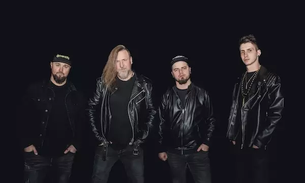 Štyria muži v čiernych kožených bundách stoja na tmavom pozadí a vyžarujú z nich vážnu a drsnú atmosféru. Niektorí majú na hlave čiapky a jeden má dlhé vlasy.