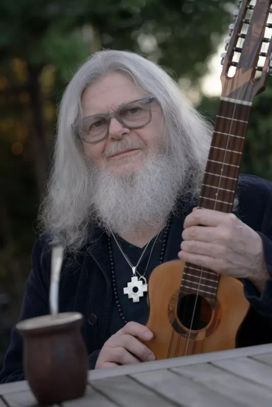 Starší človek s dlhými bielymi vlasmi a bradou, s okuliarmi a gitarou v ruke. Na drevenom stole pred ním je šálka maté, v pozadí stromy.