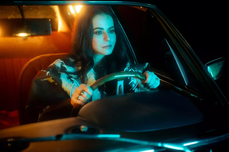 Žena s dlhými tmavými vlasmi, oblečená vo vzorovanej blúzke, sa počas jazdy v noci sústredí. Teplé vnútorné osvetlenie kontrastuje s chladným, tmavým exteriérom.