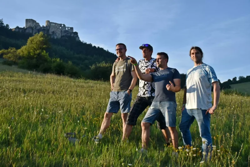 Štyria muži stoja na trávnatom kopci v ležérnom oblečení a užívajú si daný okamih. Za nimi sa na zalesnenom kopci pod jasnou modrou oblohou týči hrad.