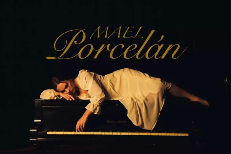 Žena v bielych šatách pokojne leží na čiernom klavíri na tmavom pozadí. Nad ňou sú elegantne zobrazené slová „Mael Porcelán“.