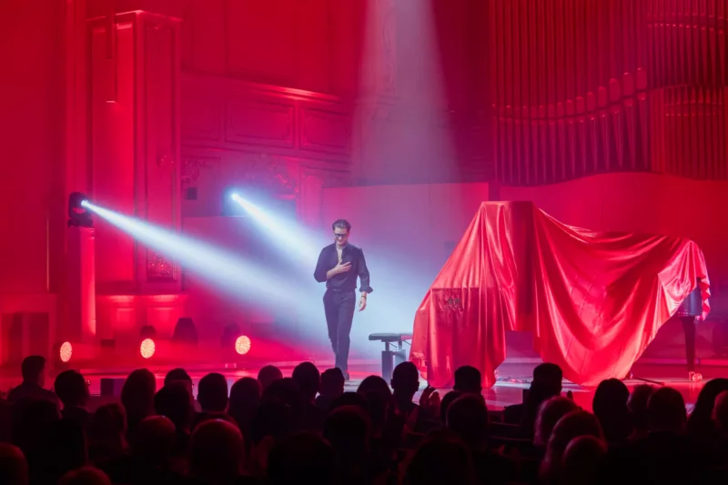 Umelec sebavedomo kráča po pódiu osvetlenom dramatickou červenou farbou s klavírom potiahnutým červenou látkou. Publikum ho dychtivo sleduje a vytvára intenzívnu a napínavú atmosféru.