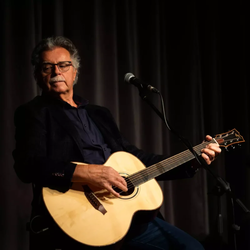 Starší muž s okuliarmi hrá na akustickej gitare na pódiu pod tlmeným svetlom. Má na sebe tmavú bundu, ktorá vyjadruje pokojnú a introspektívnu náladu.