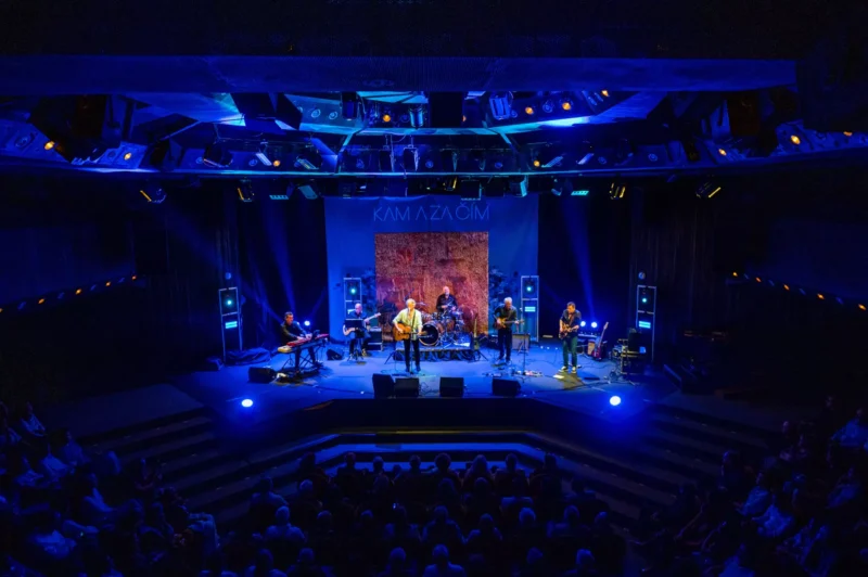 Kapela vystupuje na slabo osvetlenom pódiu s modrými a fialovými svetlami, čím vytvára živú atmosféru. Publikum sleduje vystúpenie v zatemnenom divadle.