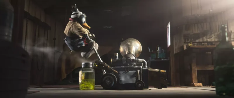 Kačica s prilbou obsluhuje stroj v slabo osvetlenej dielni. Neďaleko stojí nádoba so zelenou tekutinou. Prostredie pôsobí svojrázne a nápadito.