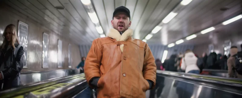 Muž v hnedej ovčej bunde a čiapke stojí na eskalátore v rušnej stanici metra a vyzerá odhodlane. V pozadí sú ľudia a rozmazané svetlá.