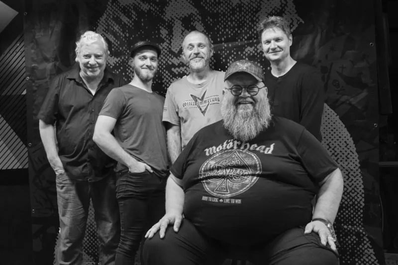 Skupina piatich mužov ležérne pózuje pred textúrovaným pozadím. Muž vpredu sedí s tričkom Motörhead, zatiaľ čo ostatní stoja za ním. Všetci sa vrúcne usmievajú a vytvárajú priateľskú a uvoľnenú atmosféru. Obrázok je čiernobiely.