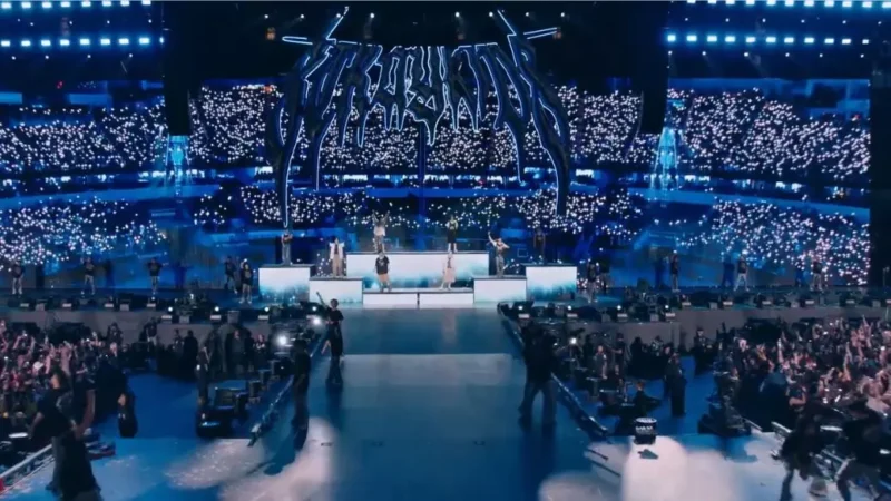 Živá koncertná scéna s kapelou vystupujúcou na jasne osvetlenom pódiu. Tisíce fanúšikov držia v zaplnenej aréne žiariace svetlá a vytvárajú tak živú atmosféru.