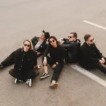 Päť ľudí v čiernom oblečení a so slnečnými okuliarmi sedí ležérne na parkovisku a vyžaruje z nich uvoľnená a cool atmosféra.