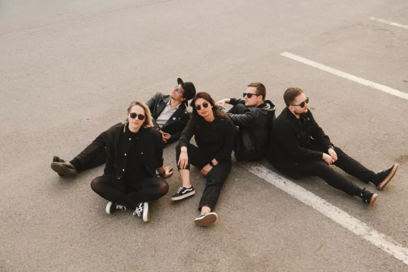 Päť ľudí v čiernom oblečení a so slnečnými okuliarmi sedí ležérne na parkovisku a vyžaruje z nich uvoľnená a cool atmosféra.