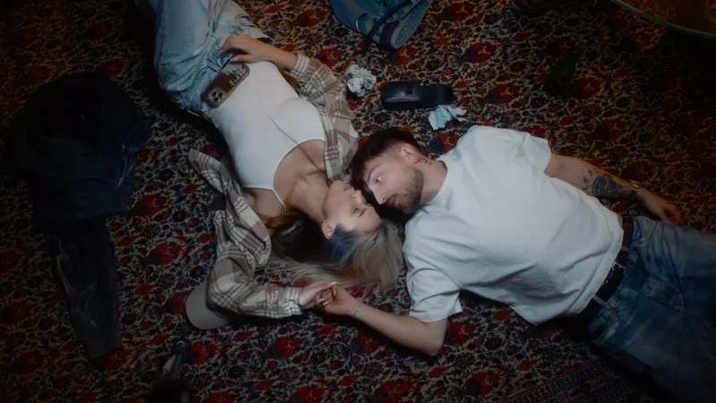 A couple lies on a patterned rug, holding hands and gazing into each other's eyes. Casual clothes, soft lighting, and scattered items create a relaxed, intimate atmosphere.