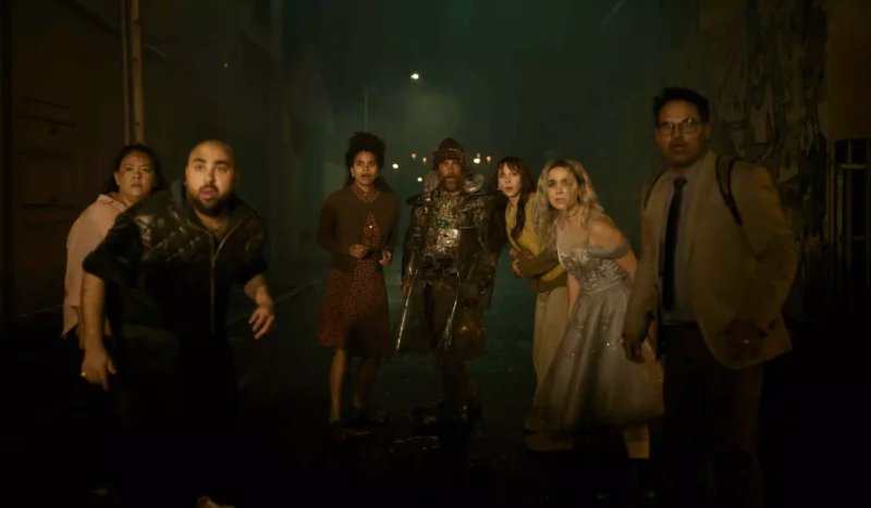 Skupina šiestich ľudí, vrátane jedného v stredovekom brnení, stojí v slabo osvetlenej uličke a s prekvapeným a znepokojeným výrazom hľadí pred seba.