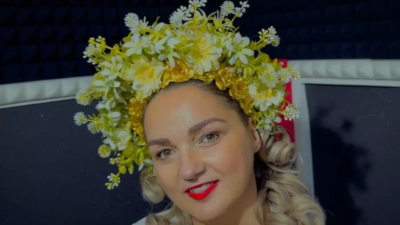 Žena so žiarivým úsmevom nosí kvetinovú korunu zo žltých a bielych kvetov. Vlasy má upravené do kučier. Pozadie tvorí tmavá stena s čalúnením.