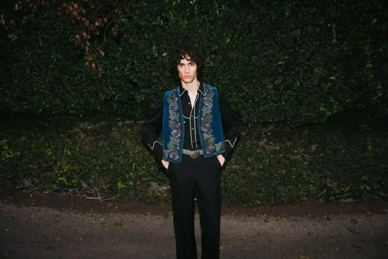 Osoba stojí pri tmavozelenom živom plote, oblečená v modrej vyšívanej veste a čiernom oblečení. Prostredie pôsobí pokojne a zamyslene.