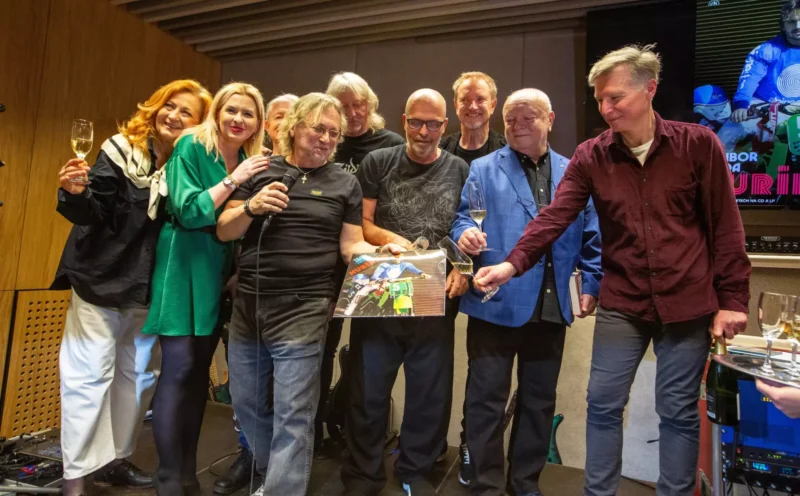 Skupina deviatich ľudí sa usmieva a oslavuje v interiéri, držiac poháre na šampanské a farebný plagát. Atmosféra je radostná a slávnostná.