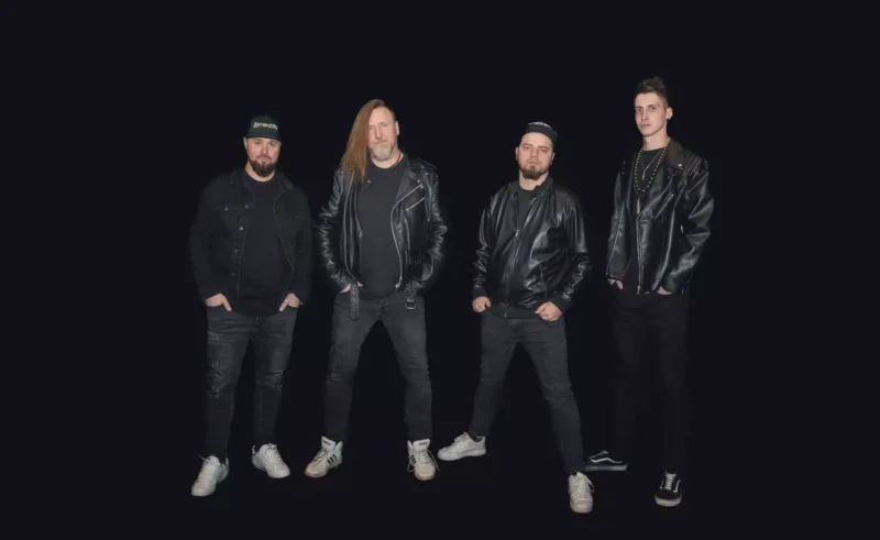 Na čiernom pozadí stoja štyria muži, všetci majú na sebe čierne bundy, tmavé nohavice a tenisky. Traja majú fúzy, jeden má dlhé vlasy a pózujú s rukami vo vreckách alebo po bokoch.