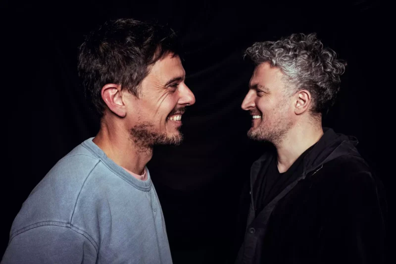 Dvaja muži stoja tesne oproti sebe na tmavom pozadí a obaja sa vrúcne usmievajú. Vyžaruje z nich priateľstvo a kamarátstvo.