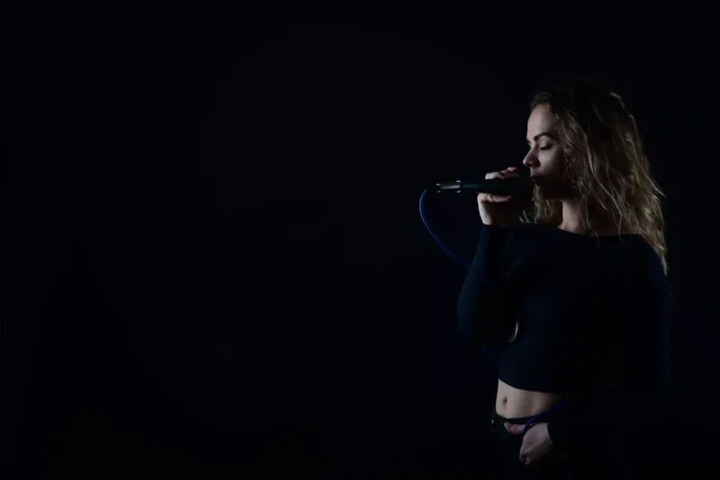 Žena v slabo osvetlenom prostredí spieva do mikrofónu so zatvorenými očami, pričom vyžaruje pokojnú a sústredenú náladu. Má vlnité vlasy a tmavý top.