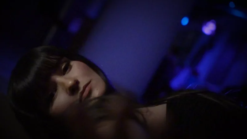 A woman with long dark hair gazes introspectively in a dimly lit room with blue and purple hues, creating a moody and contemplative atmosphere.