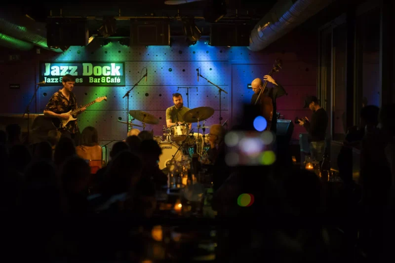 Džezová kapela vystupuje na pódiu v Jazz Docku s gitarou, bicími a kontrabasom pod farebnými svetlami. Publikum počúva v tlmenom, útulnom prostredí.