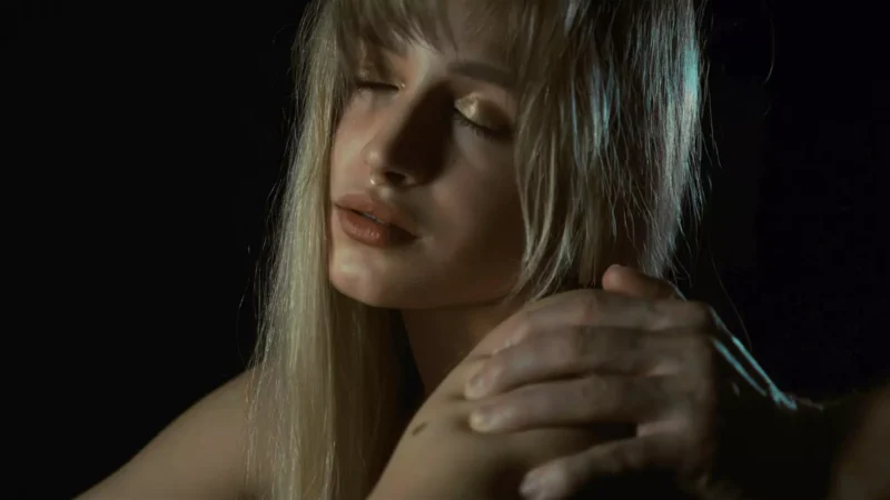 Žena s dlhými blond vlasmi zatvára oči, keď jej na tmavom pozadí jemne spočíva ruka na ramene.