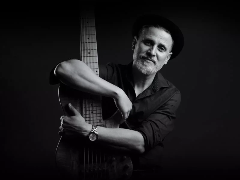 Čiernobiely portrét usmievavého muža s bradou, ktorý má na sebe klobúk a hodinky a drží v rukách akustickú gitaru. Nálada je uvoľnená a umelecká.