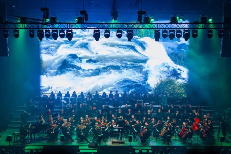 Orchester vystupuje na pódiu s veľkou osvetlenou obrazovkou za ním, ktorá zobrazuje dynamickú scénu vodopádu. Hudobníci hrajú sústredene a vášnivo.