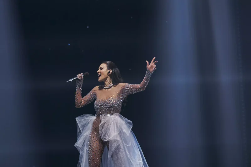 Účinkujúca v trblietavých, priehľadných šatách vášnivo spieva na pódiu. Drží mikrofón a okolo nej vrhá dramatické svetlo lúče.