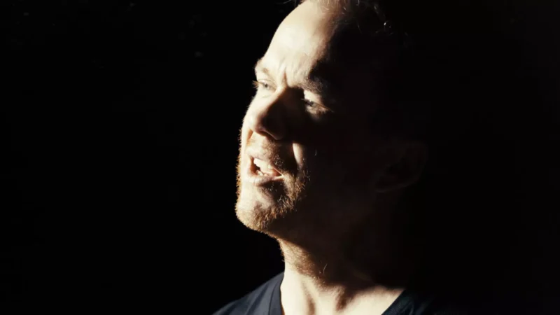 Muž je osvetlený dramatickým bočným svetlom na tmavom pozadí. Má zatvorené oči a zdá sa, akoby spieval, čím vyjadruje emócie a intenzitu.