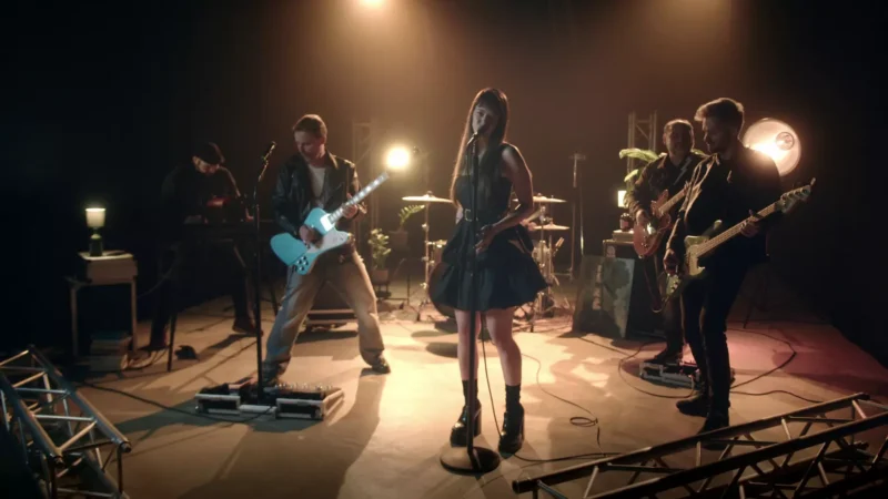 Kapela energicky vystupuje na pódiu, v ktorej účinkuje speváčka v čiernych šatách a štyria hudobníci s gitarami a bicími pod dramatickým osvetlením.