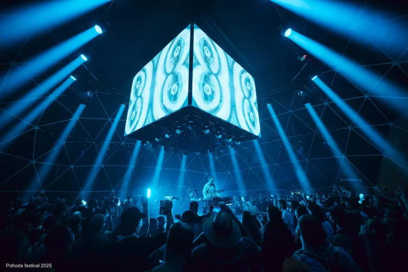 Živá koncertná scéna s kockou zobrazujúcou abstraktné vizuály zavesené nad ňou. Modré svetlá osvetľujú jásajúci dav a vytvárajú energickú atmosféru.