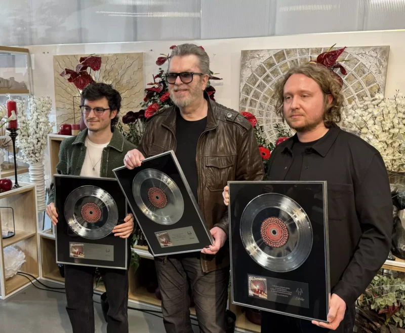Traja muži držia strieborné platne s platňami a stoja pred kvetinovou výzdobou a umeleckými dielami. Nálada je slávnostná a hrdá.