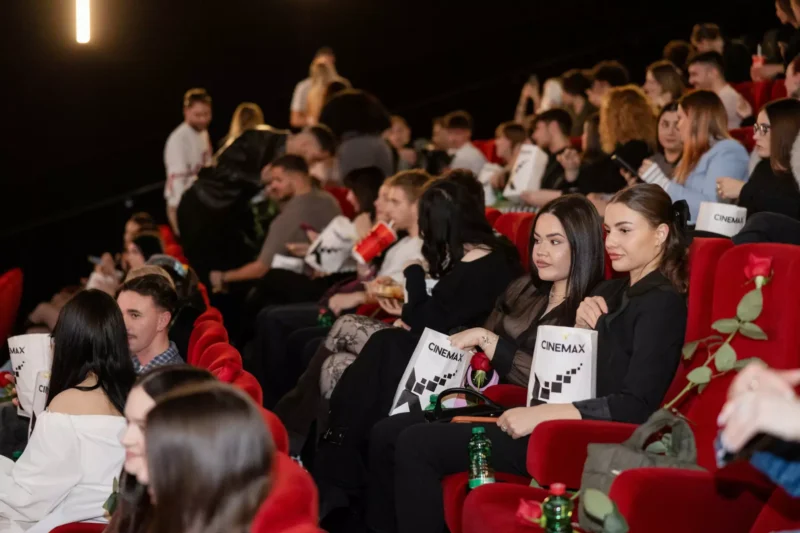 Diváci sedia v slabo osvetlenom kine a držia v rukách pukance a nápoje. Ľudia vyzerajú zaujato a nadšene. Viditeľné sú jasne červené sedadlá a balené občerstvenie.