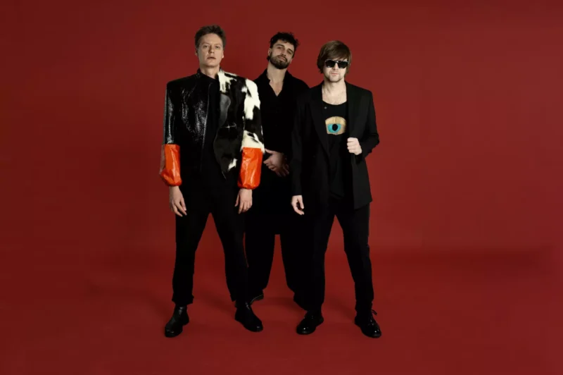 Traja muži stoja pred červeným pozadím. Jeden má na sebe výraznú bundu s oranžovými rukávmi, ostatní v čiernej, jeden so slnečnými okuliarmi, čo vytvára drzú a sebavedomú atmosféru.