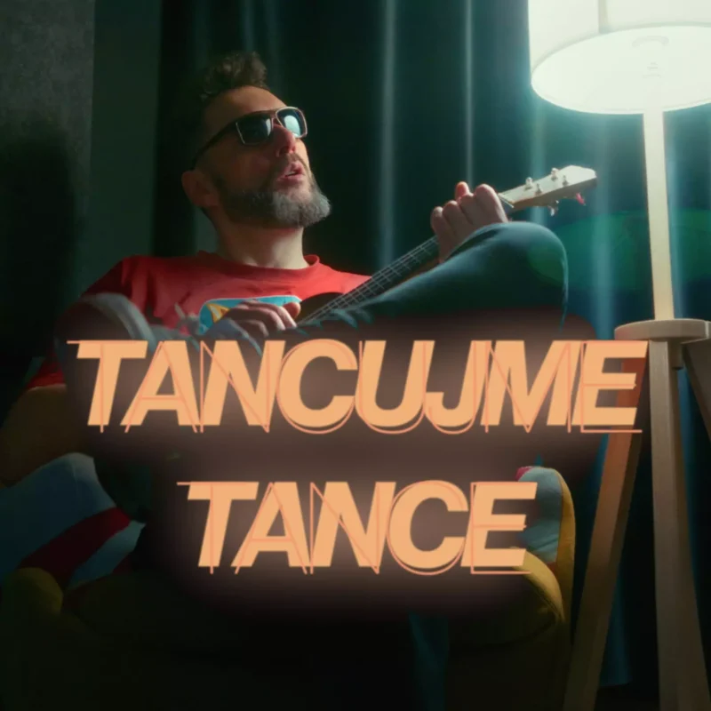 Muž v slnečných okuliaroch hrá na gitare v tmavej miestnosti, sedí vedľa stojacej lampy. Tučný text v popredí znie „TANCUJME TANCE“, čo vytvára uvoľnenú atmosféru.