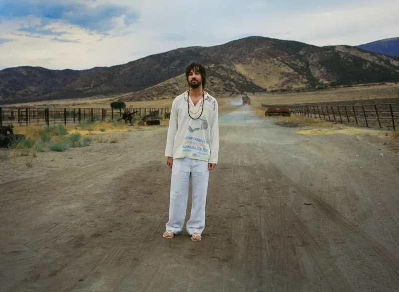 Osoba v ležérnom oblečení stojí na prašnej ceste v rozsiahlej, vyprahnutej krajine, lemovanej horami a plotmi, a evokuje pocit samoty.