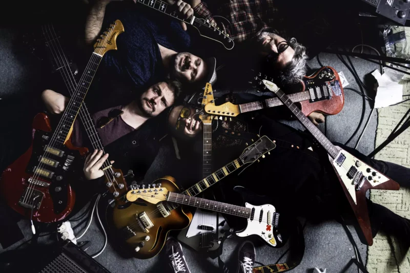 Štyria ľudia ležia na podlahe, obklopení rôznymi gitarami. Scéna vyvoláva uvoľnenú, kreatívnu atmosféru a zdôrazňuje vášeň pre hudbu.