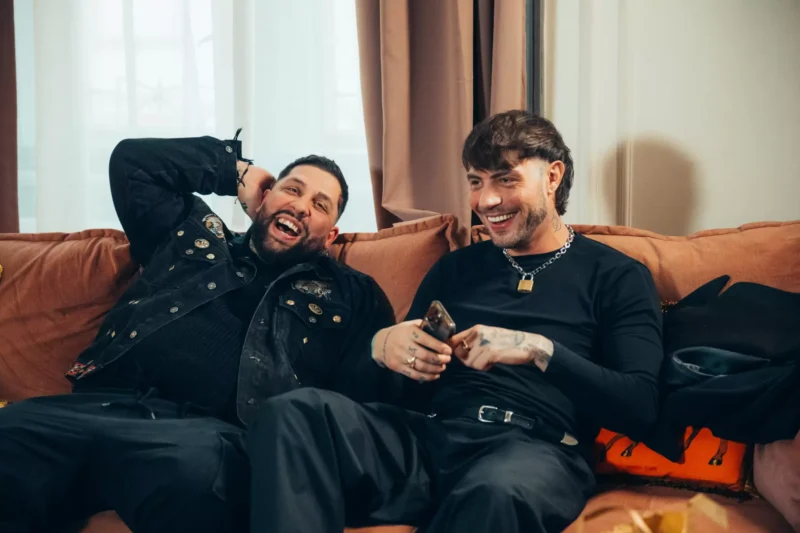 Dvaja ľudia sedia na hnedej pohovke a smejú sa spolu. Jeden má na sebe čiernu bundu a druhý drží telefón. Atmosféra je uvoľnená a radostná.