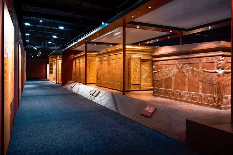 Slabo osvetlená múzejná expozícia s veľkým, zdobeným egyptským sarkofágom s vyrytými hieroglyfmi, pod ostrým svetlom reflektorov. Tajomná a historická atmosféra.